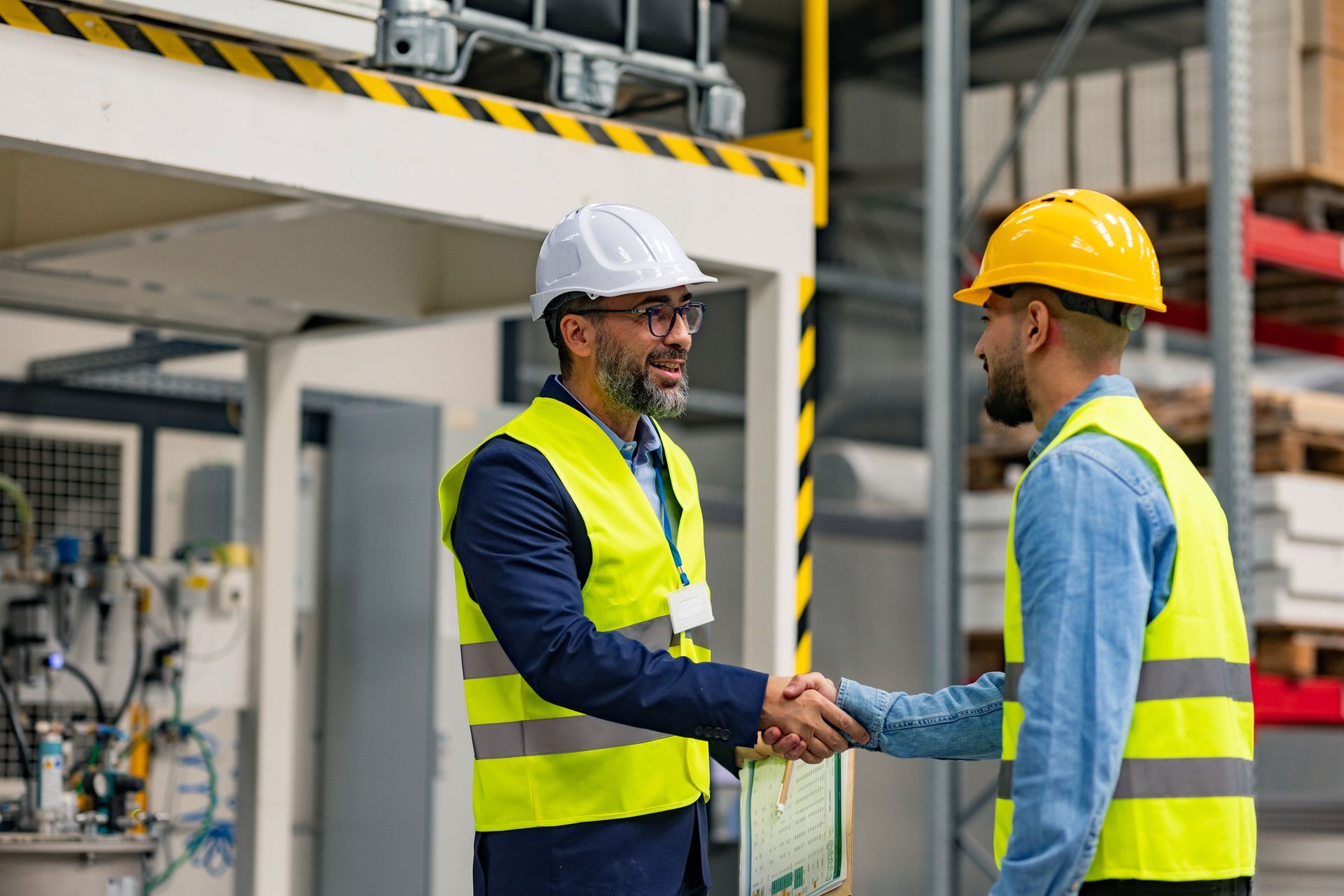 Two men in safety vests and hard hats shaking hands in a factory setting.