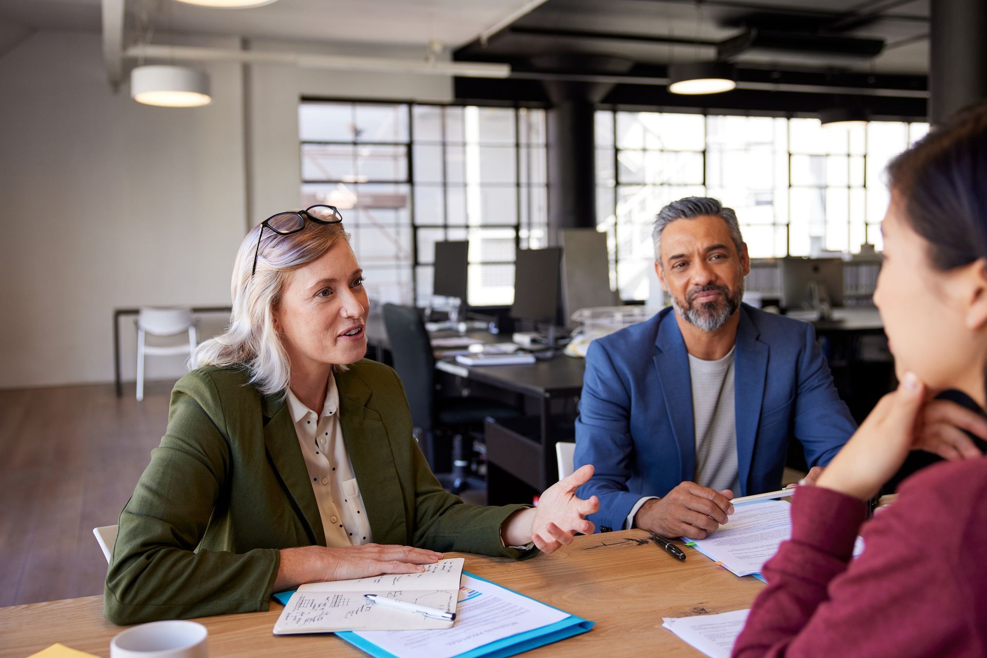 Three people in a meeting; a woman in green jacket gestures while speaking, two others listen. Office setting.