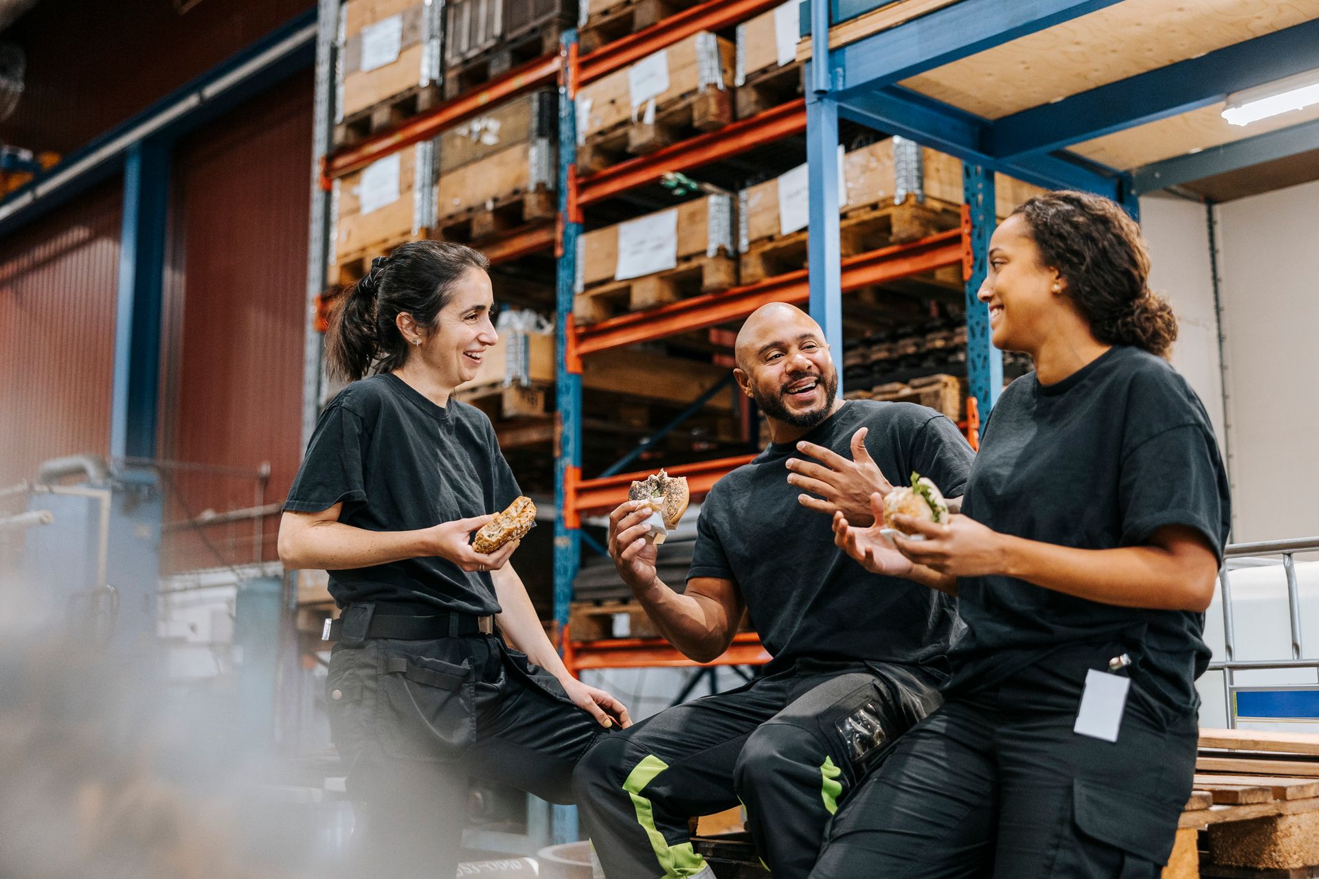 Three warehouse workers taking a lunch break, laughing, eating sandwiches in front of storage racks.
