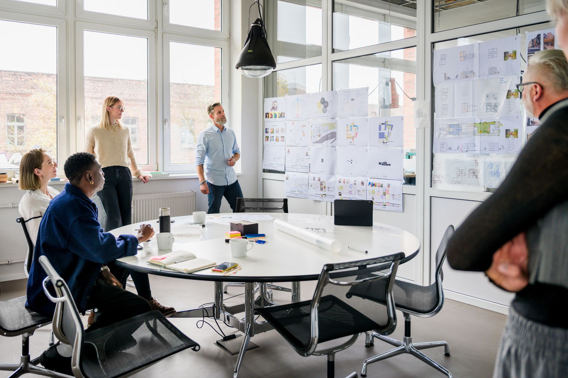People in an office meeting, looking at papers on a glass wall. Light-filled room with a circular table.