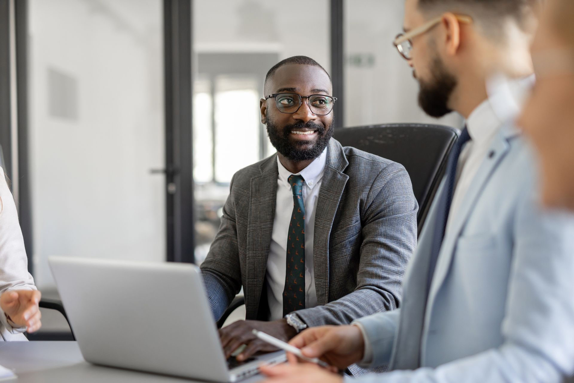 Black man in a grey suit smiles while looking at a laptop during a meeting with coworkers in an office.