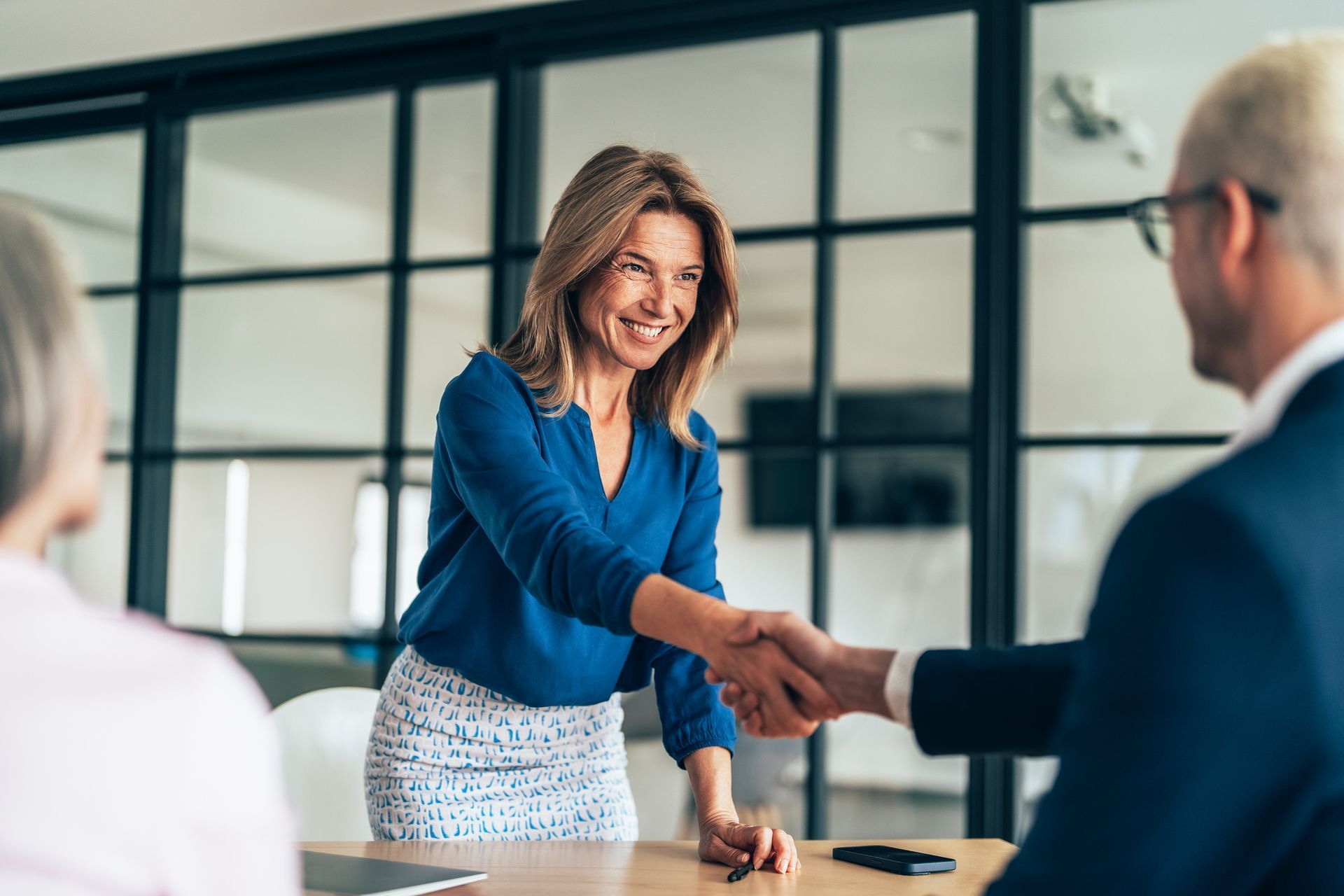 Woman in blue shirt shakes hands with a man in a suit, smiling, in an office setting.