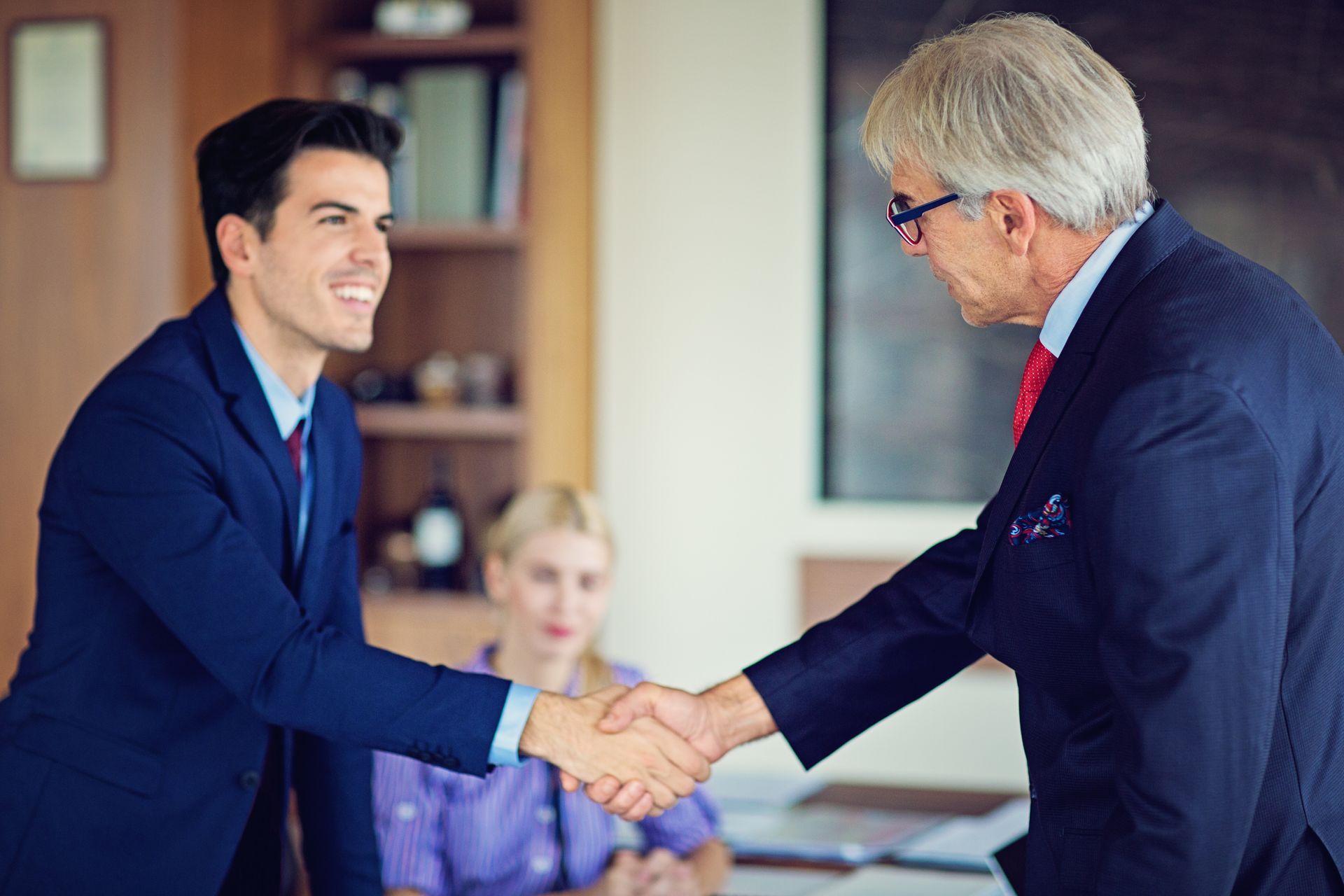 Man in blue suit shakes hands with older man in a suit; a woman watches in the background.