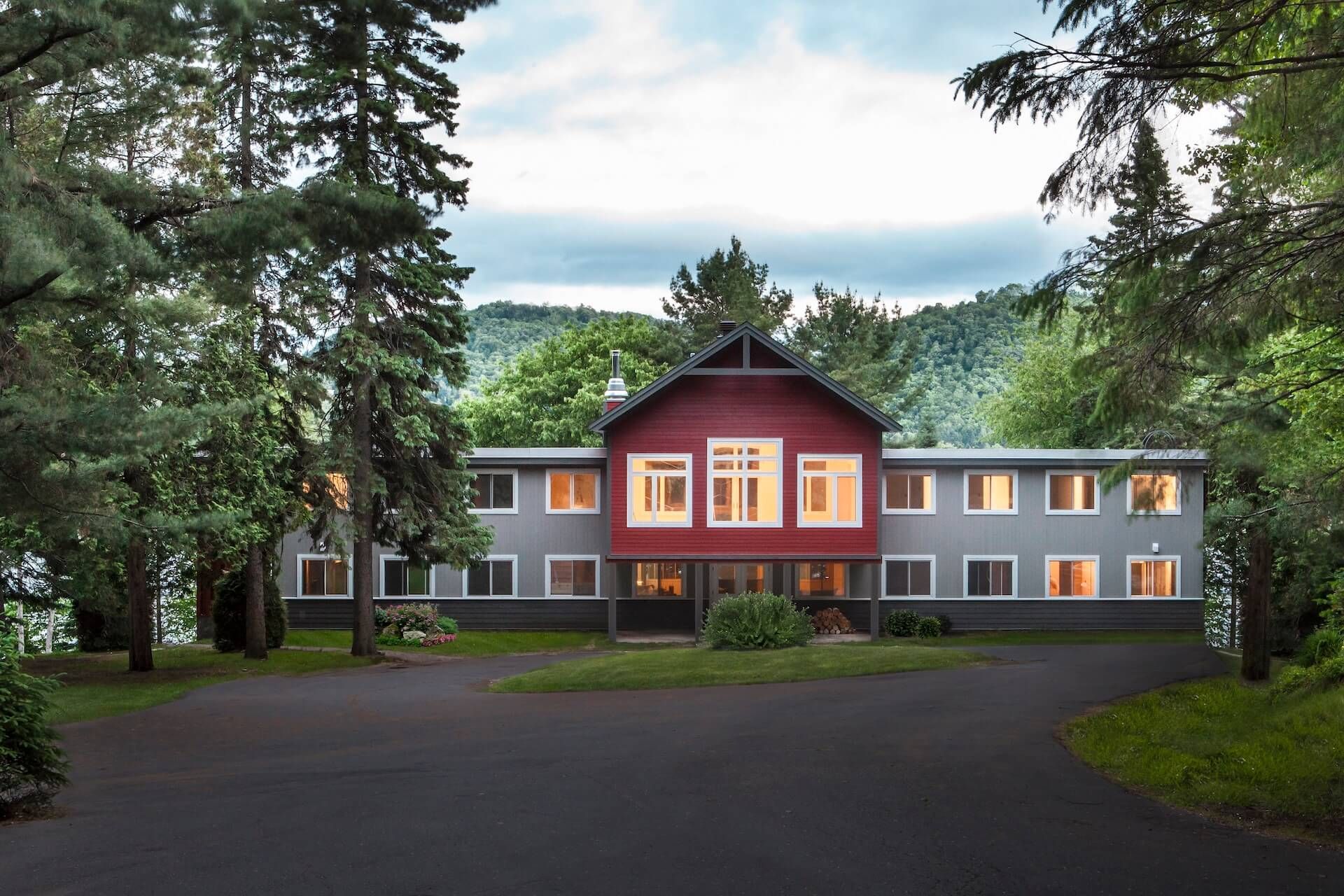 Gray and red lodge nestled in a forest, lit windows, dark driveway, trees in foreground and background.