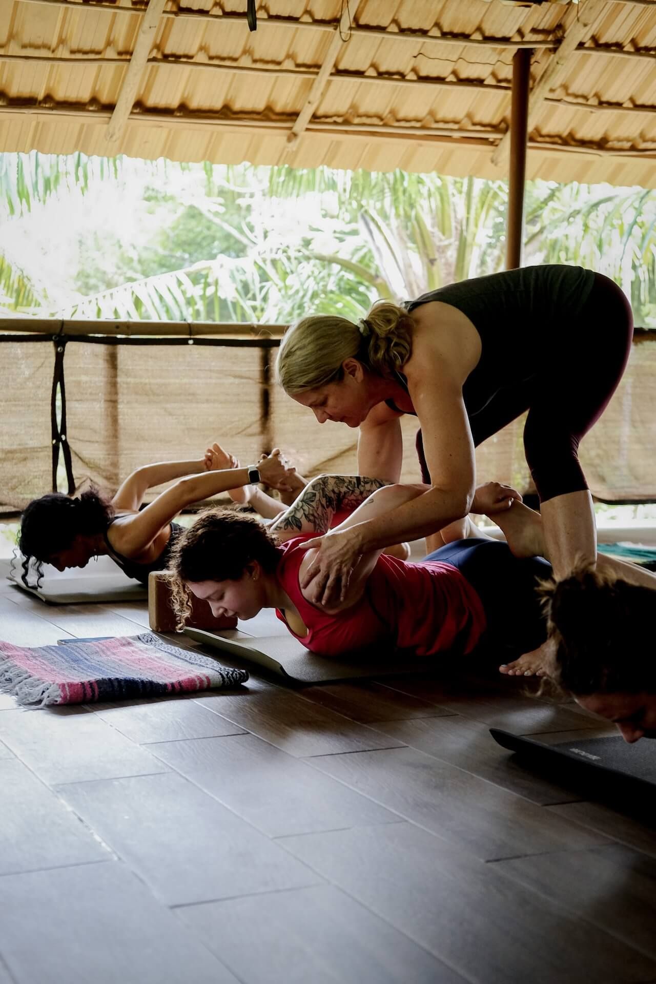 Yoga instructor assisting students in a backbend pose indoors with a wooden floor and bamboo roof.