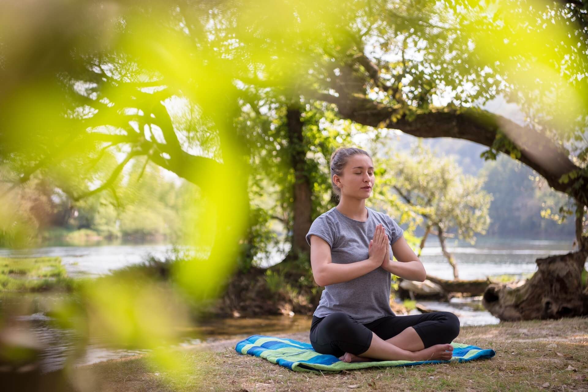 A woman is sitting in a lotus position on a yoga mat in the woods.
