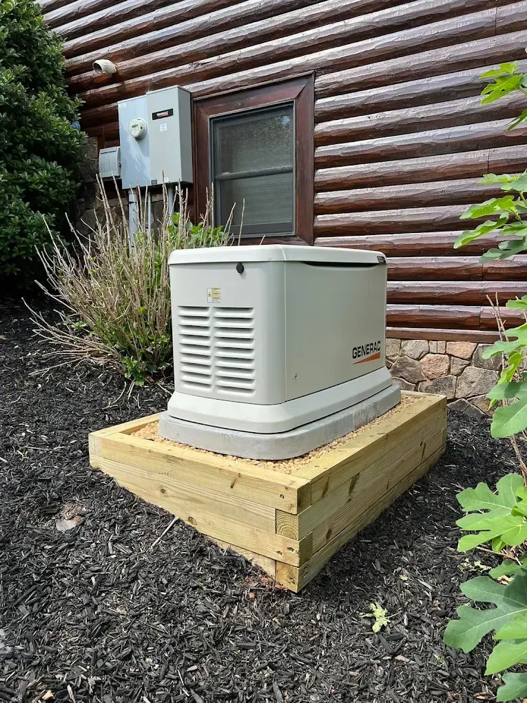Generator on a wooden platform next to a log cabin. Gray generator, wooden platform, mulch, and bushes.