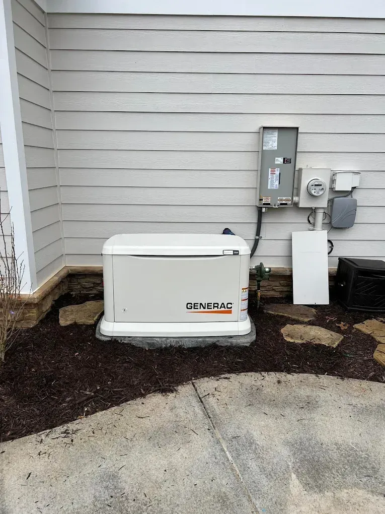 A standby generator on a concrete pad next to a house with electrical components. Ground covered in mulch.