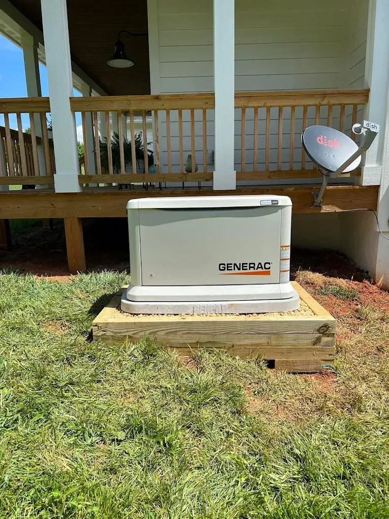 A Generac home generator is mounted on a wooden platform near a house with a deck and satellite dish.