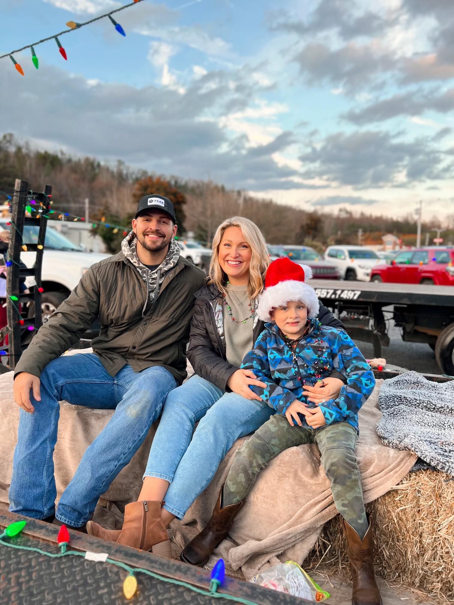 Family sitting outdoors, wearing warm clothes. Child in Santa hat, smiles. Strings of lights overhead.