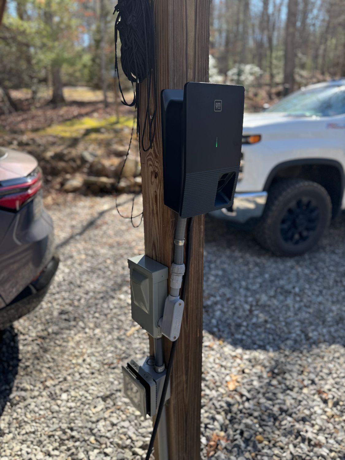 Black EV charger attached to a wooden pole, with wiring and a gray electrical box. A truck is in the background.