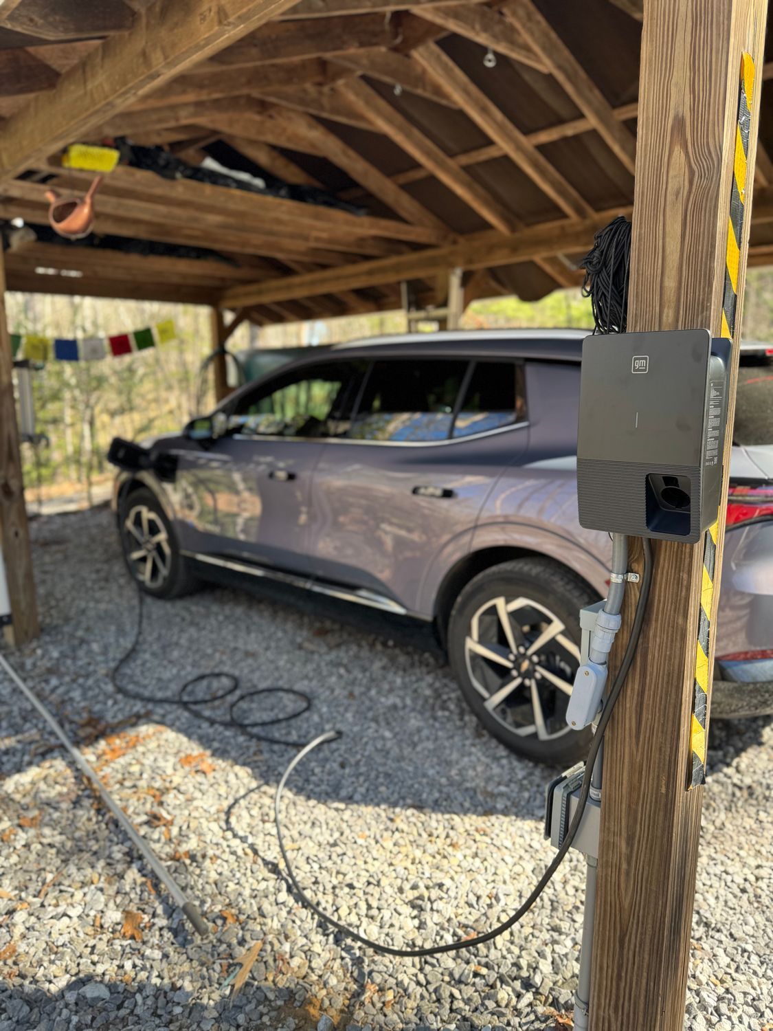 Electric vehicle charging under a wooden shelter, with charging cable connected. Gray car parked on gravel.