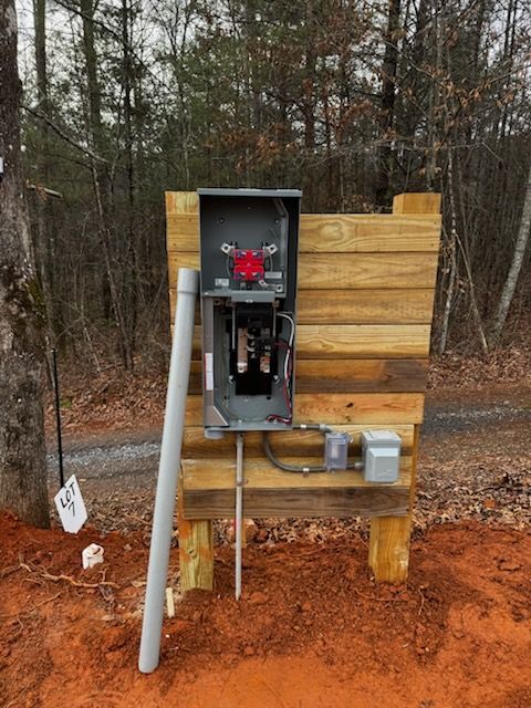 Electrical panel mounted on a wooden frame, conduit, and buried wire in a dirt clearing near trees.