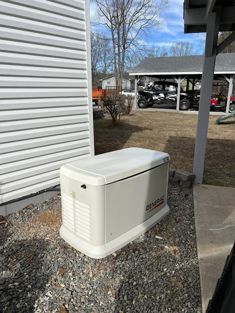 Beige generator on gravel beside a house with white siding and a carport in the background.
