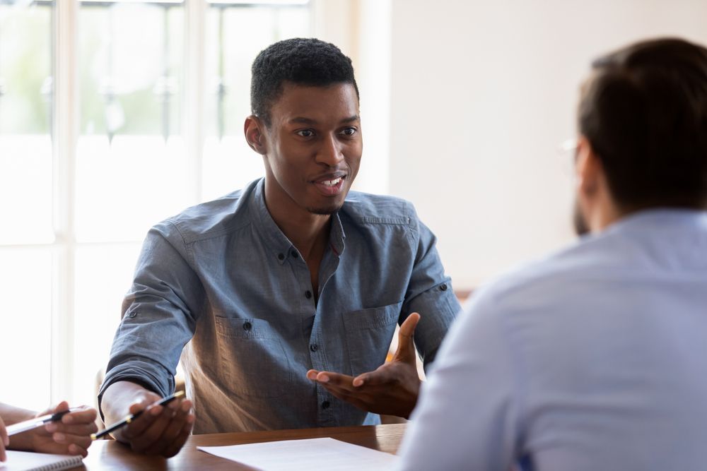 A man is sitting at a table talking to another man.