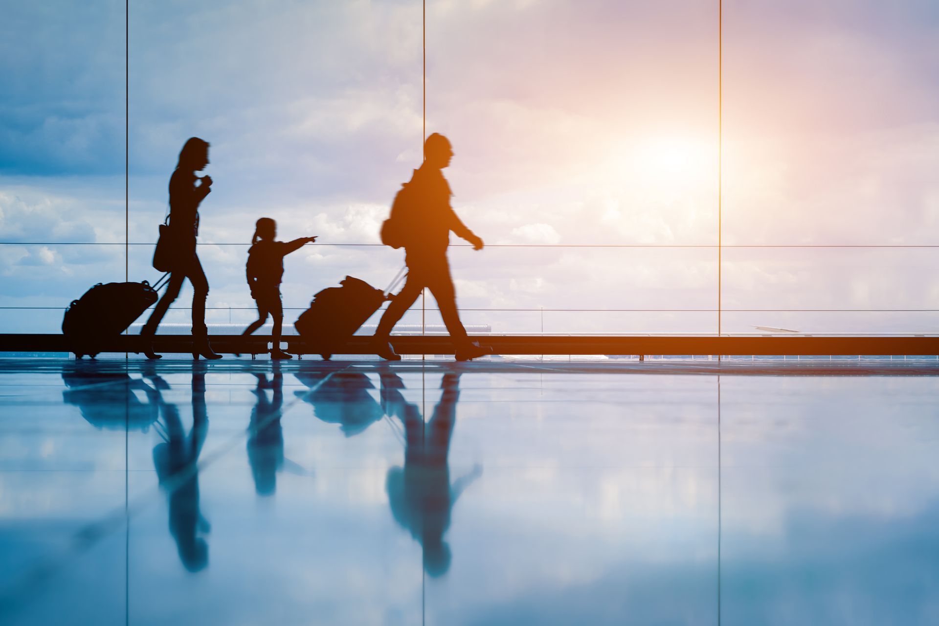 Silhouetted travelers with luggage walking through an airport at sunrise, reflected on the floor