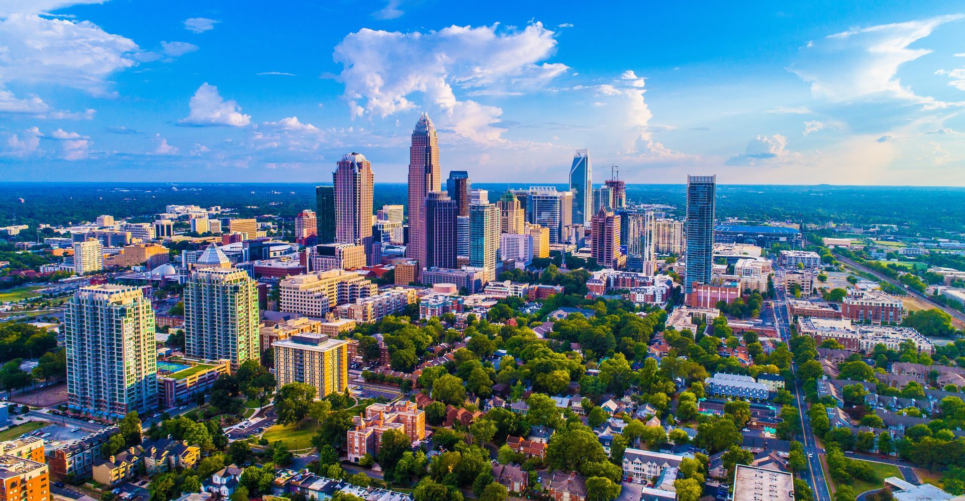 Aerial view of a city skyline with tall buildings, green neighborhoods, and a bright blue sky with clouds.