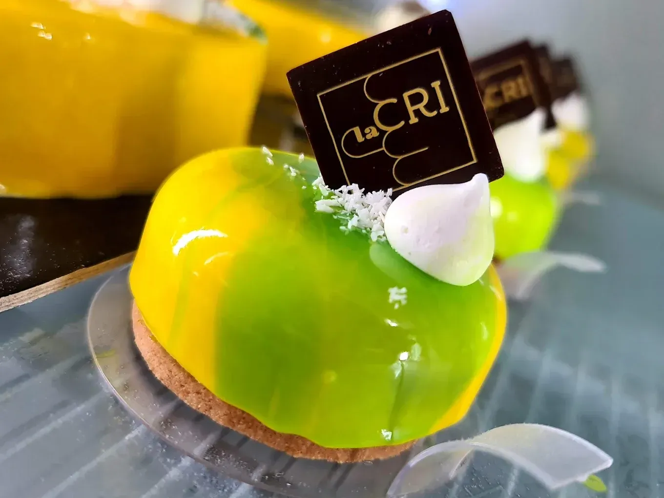 Dessert dai colori vivaci con una glassa verde e gialla lucida, guarnito con meringa bianca e un logo di cioccolato.