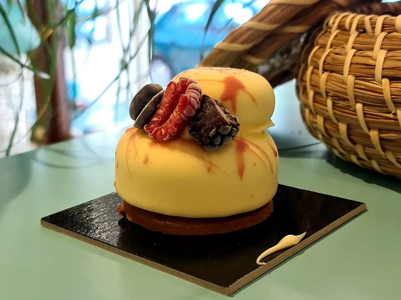 Piccolo dolcetto giallo con bacche su una base nera quadrata, con un cestino intrecciato e uno sfondo blu sfocato.