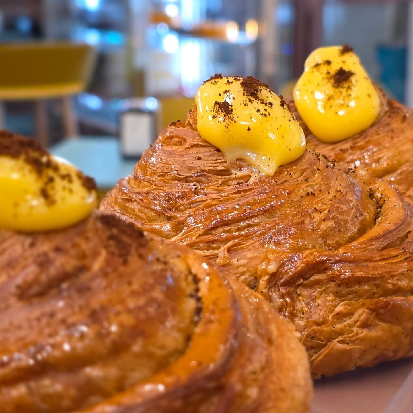 Primo piano di tre pasticcini dorati ricoperti di crema gialla e cacao in polvere, all'interno di un bar.