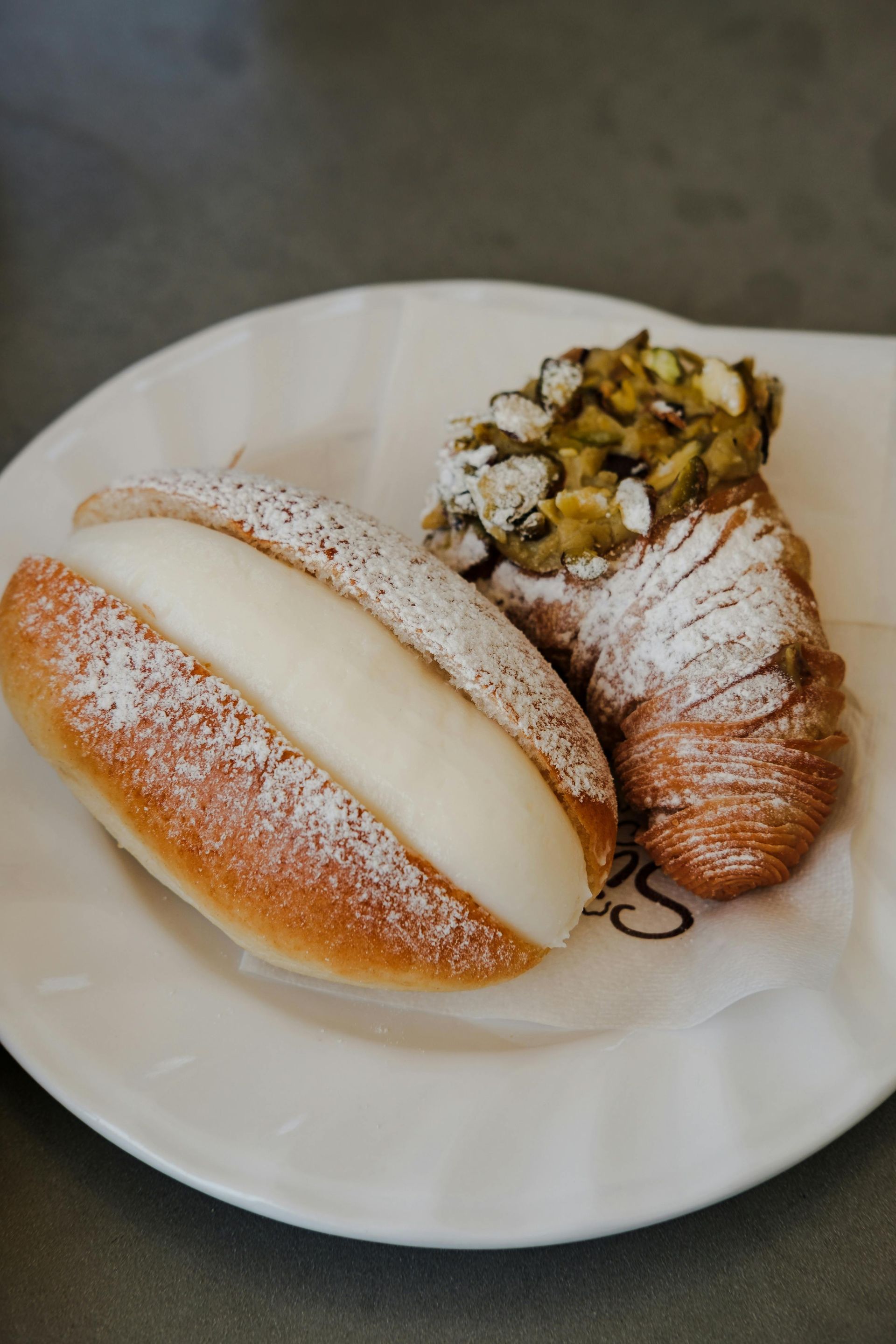 Pasticcini su un piatto bianco: panino ripieno di crema, croissant con pistacchio e un altro dolce cosparso di zucchero a velo.