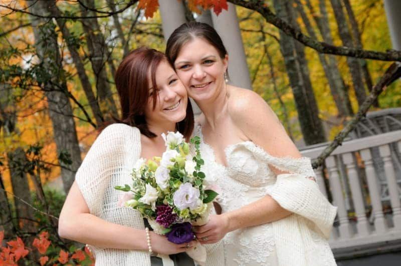 Two women in wedding dresses are posing for a picture . one of the women is holding a bouquet of flowers.
