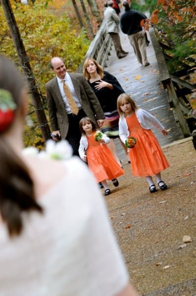 A group of little girls in orange dresses are walking across a bridge