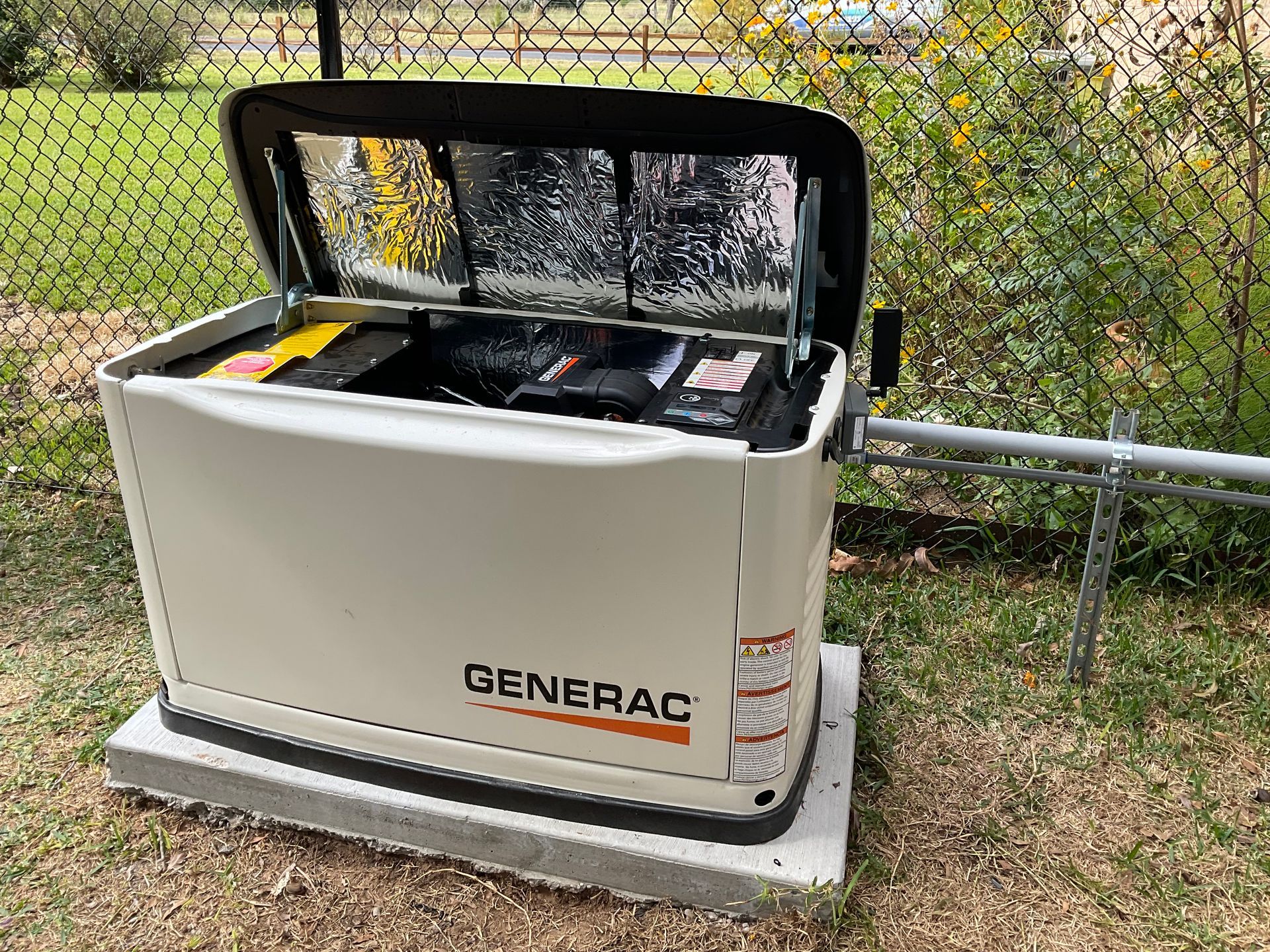 A Generac home generator, beige, open top, sitting on a concrete pad beside a chain-link fence.