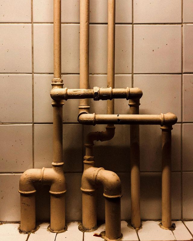 Old brass pipes and elbows mounted on a tiled wall above a concrete floor