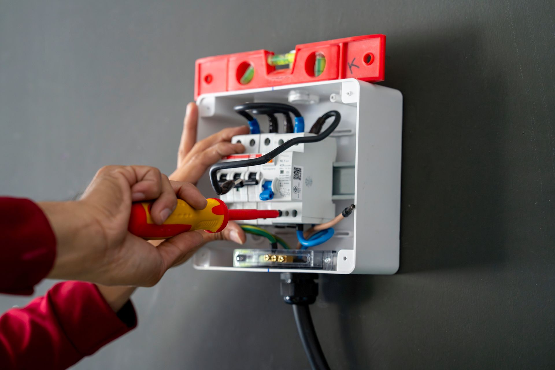 Electrician working on a white electrical box mounted on a gray wall, using a screwdriver, with a level on top.