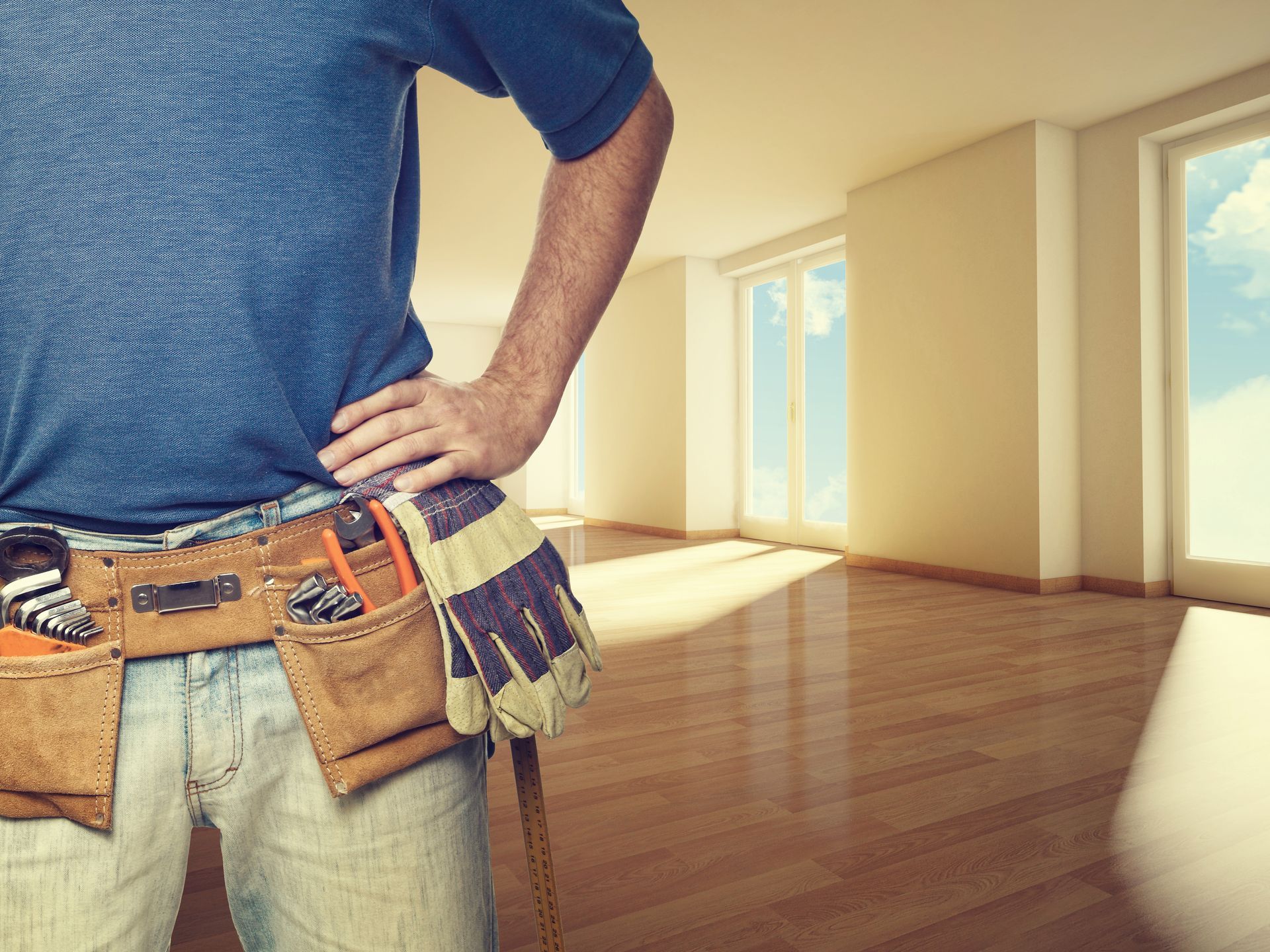 A man wearing a tool belt and gloves is standing in an empty room.