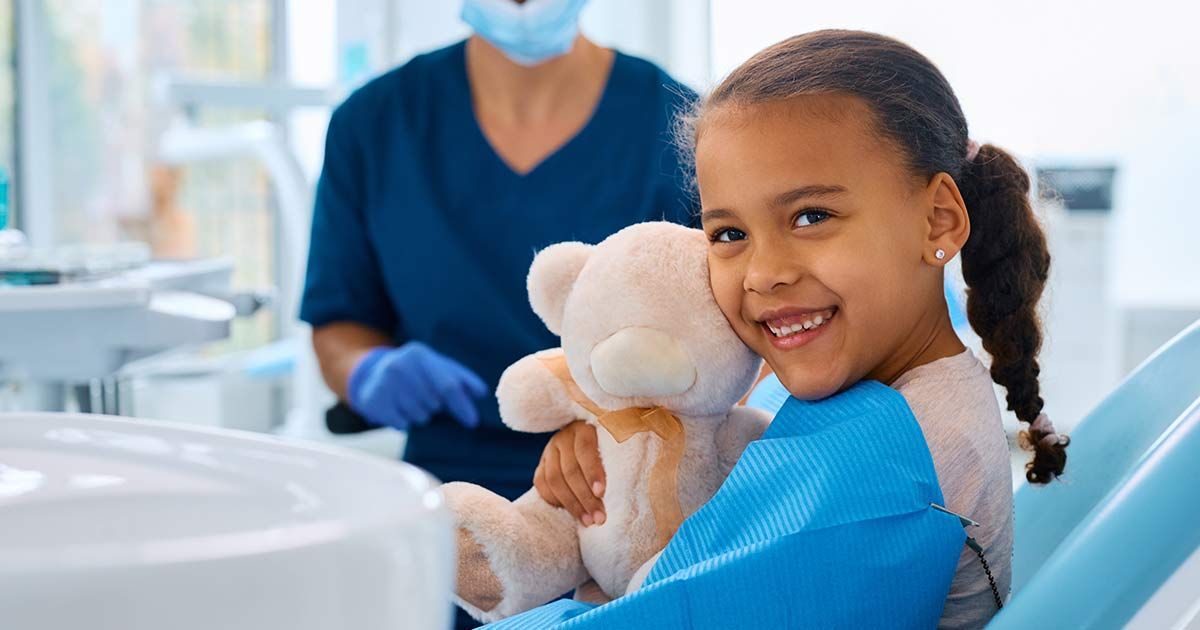 A little girl is sitting in a dental chair holding a teddy bear.