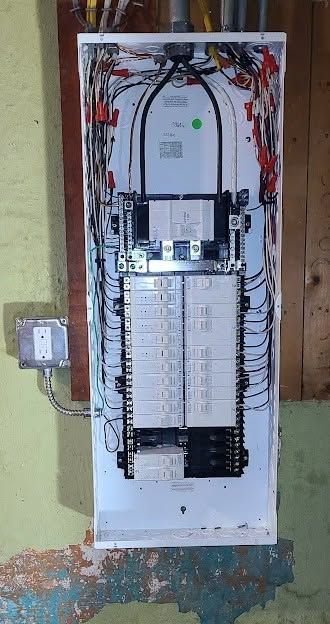 Open electrical service panel with wiring and circuit breakers mounted on a plywood wall.