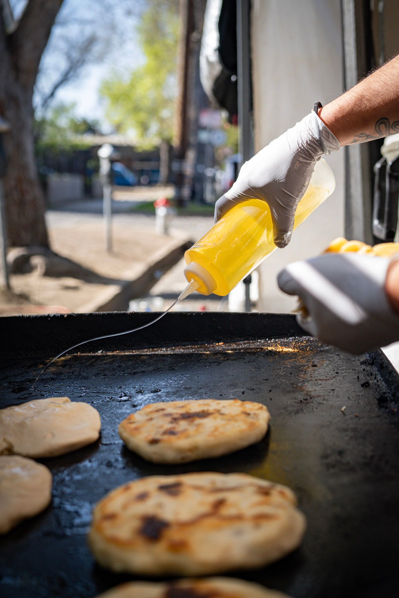 A person in gloves pouring oil on flatbreads cooking on a hot griddle outdoors.
