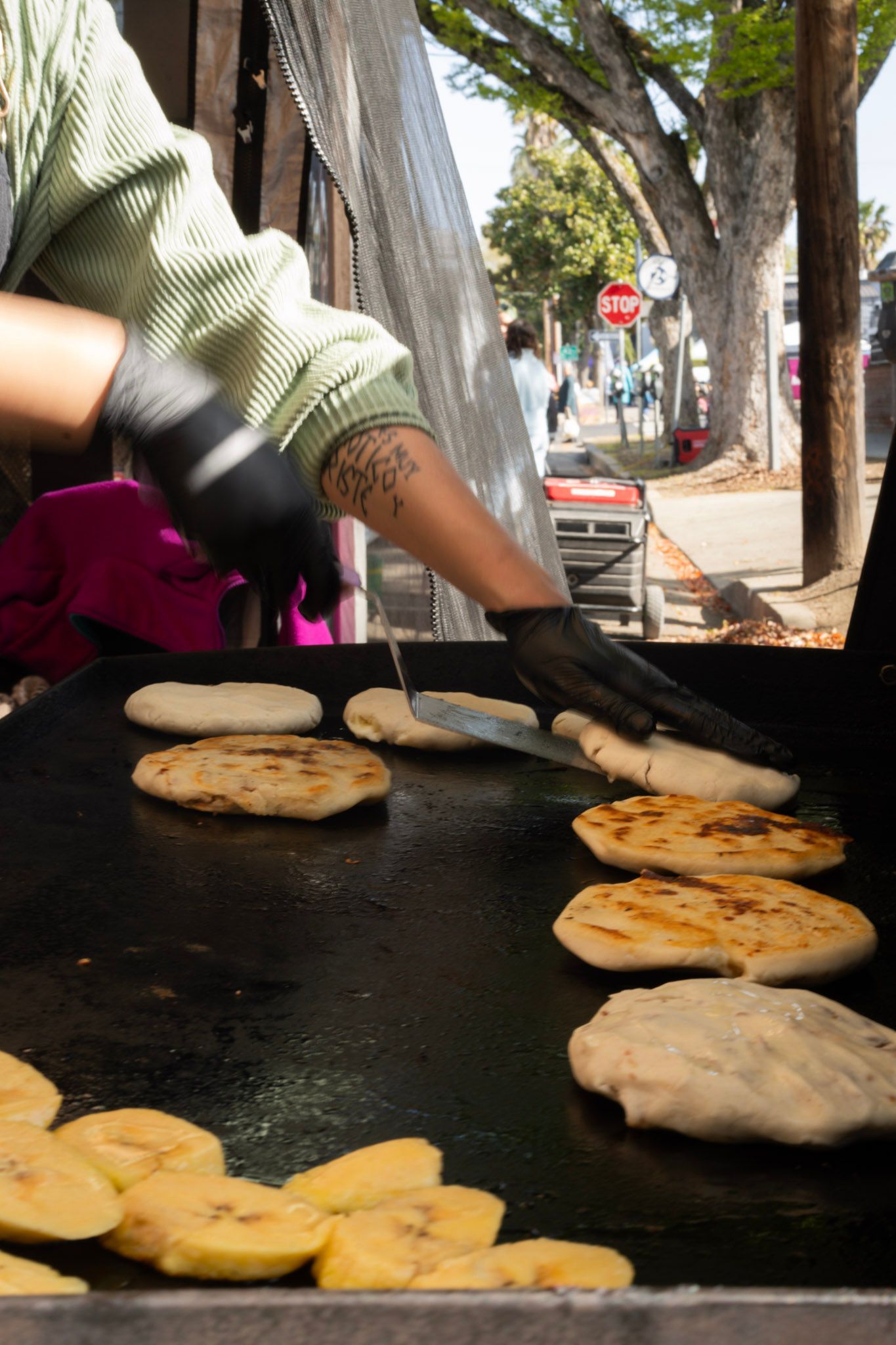 A person grilling arepas and plantains outdoors, using a spatula, sunny day.