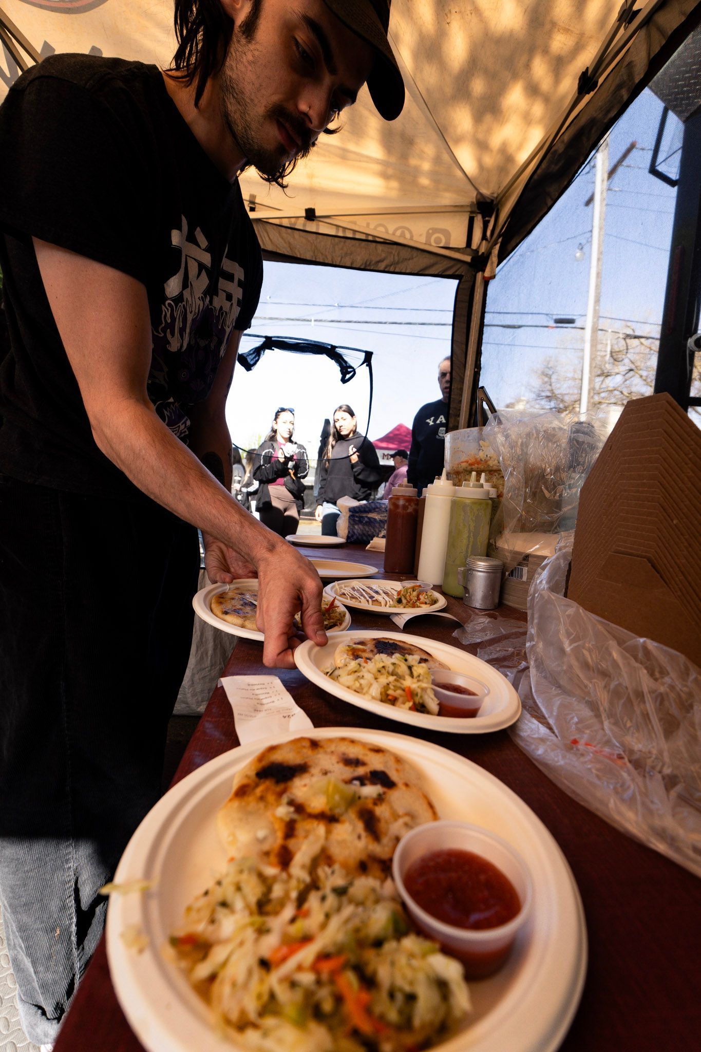 Man preparing food at a food stand. Plates of food with salsa and coleslaw visible.