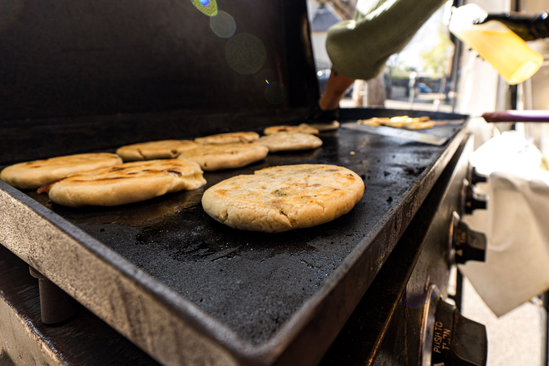 Arepas cooking on a hot griddle; cook's hand points, sunshine.