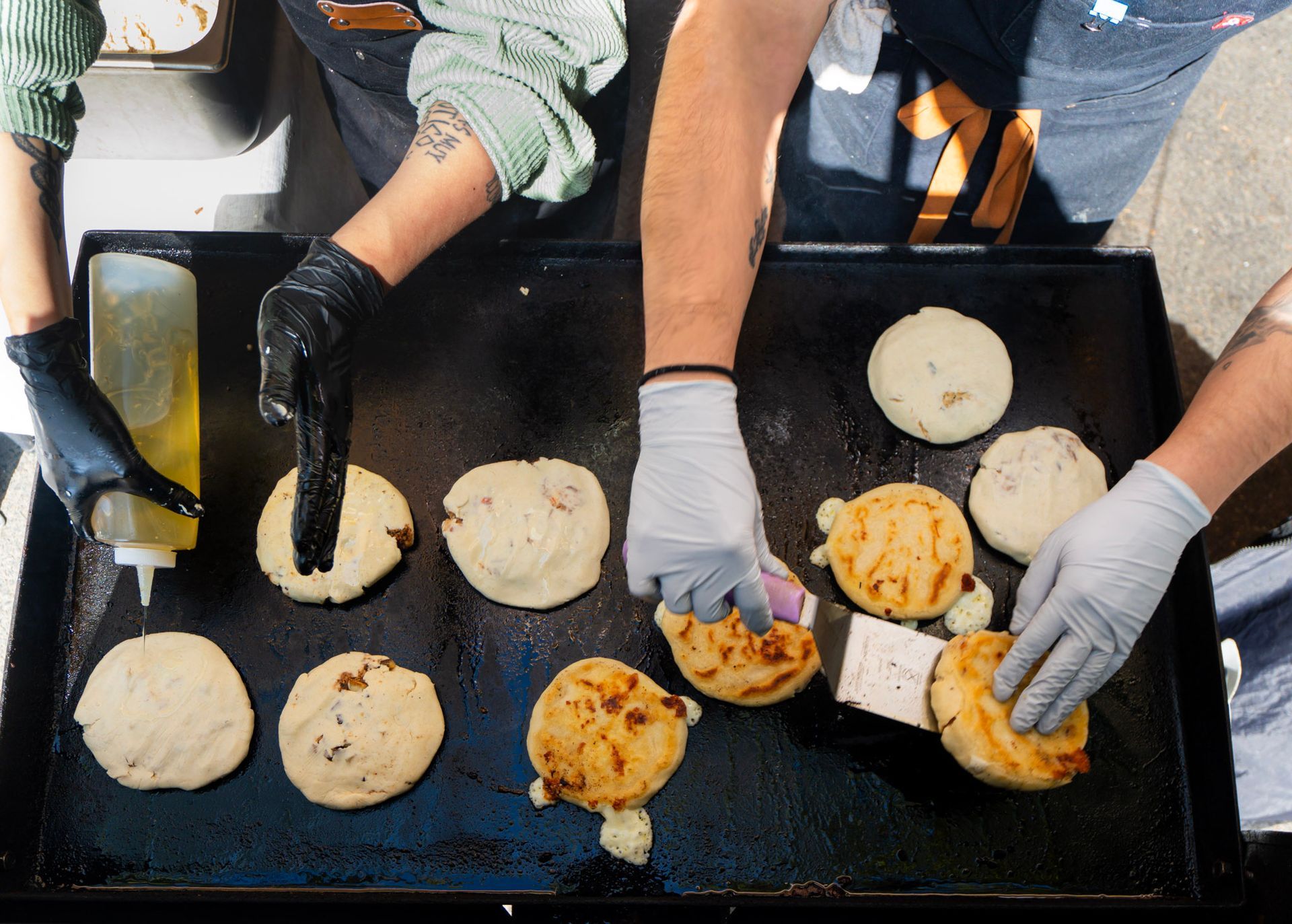Three people grilling doughy, circular arepas on a black flat top grill. One person adds oil, another flips them.