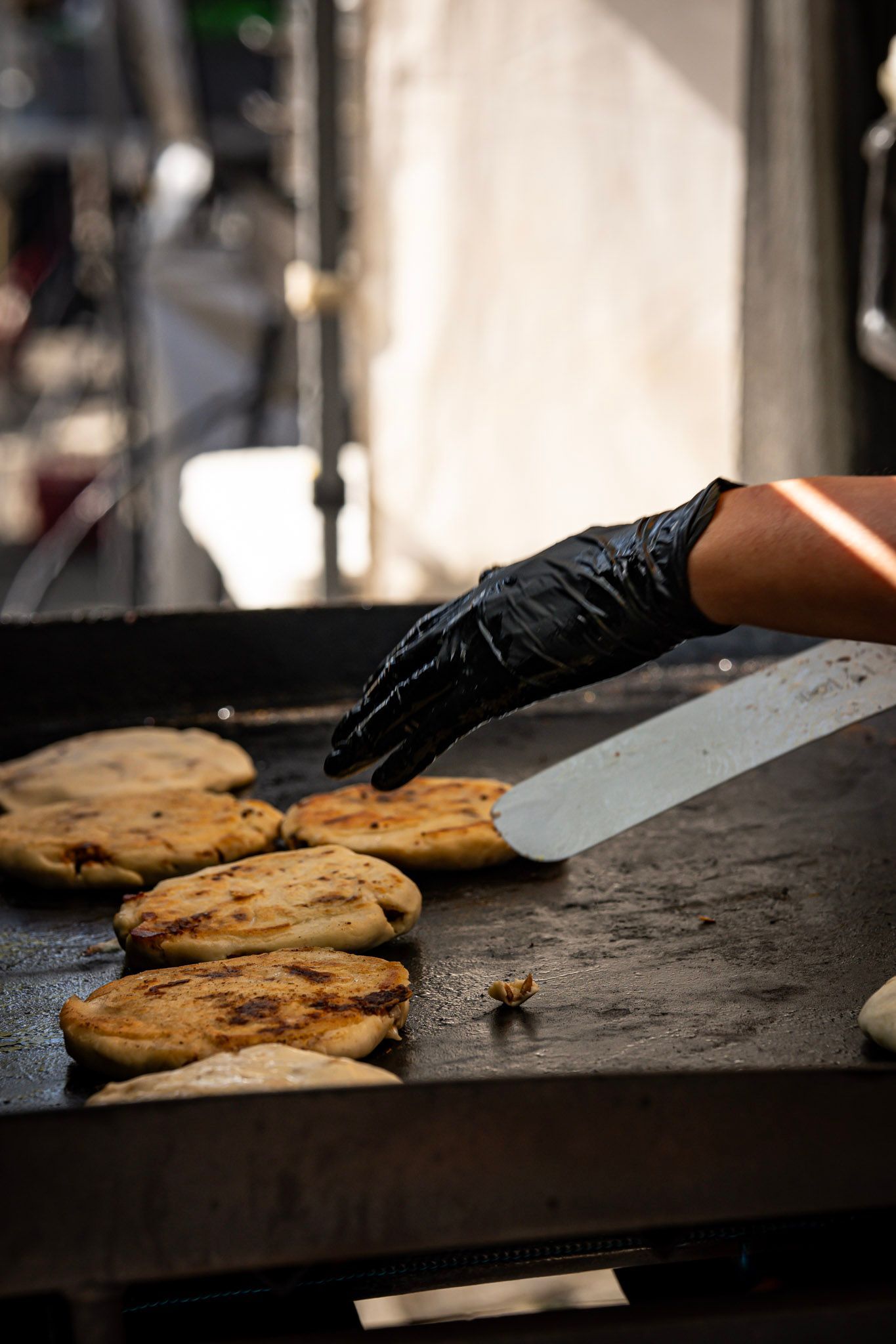 A person in black gloves uses a spatula to flip arepas on a hot griddle.