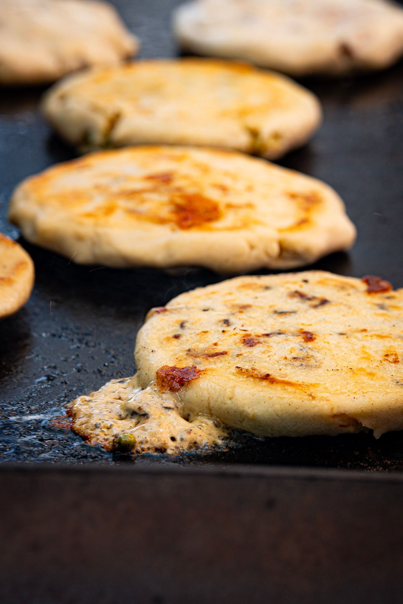 Puffy, golden-brown pupusas cooking on a flat griddle, some with cheese oozing.