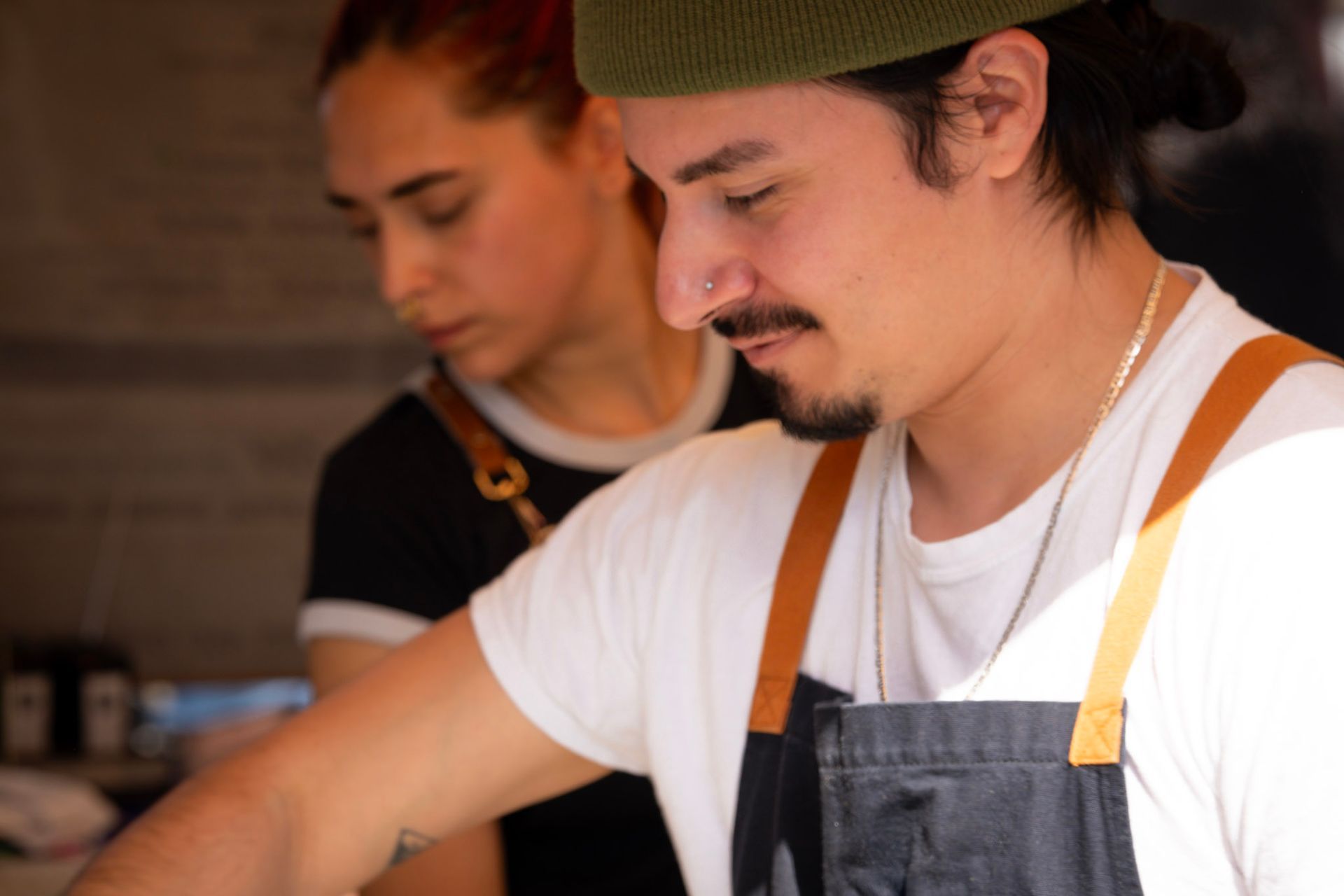 Two people working in a kitchen. Man with mustache and green hat, focused. Woman with red hair, looking down.