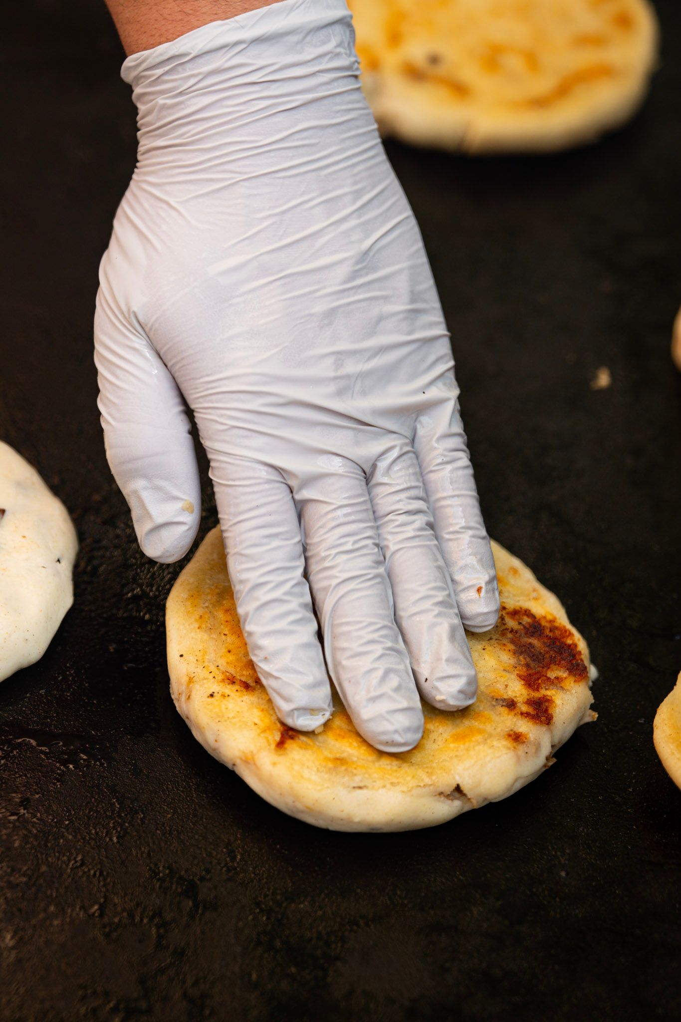 A gloved hand presses down on a round, browned flatbread cooking on a dark griddle.