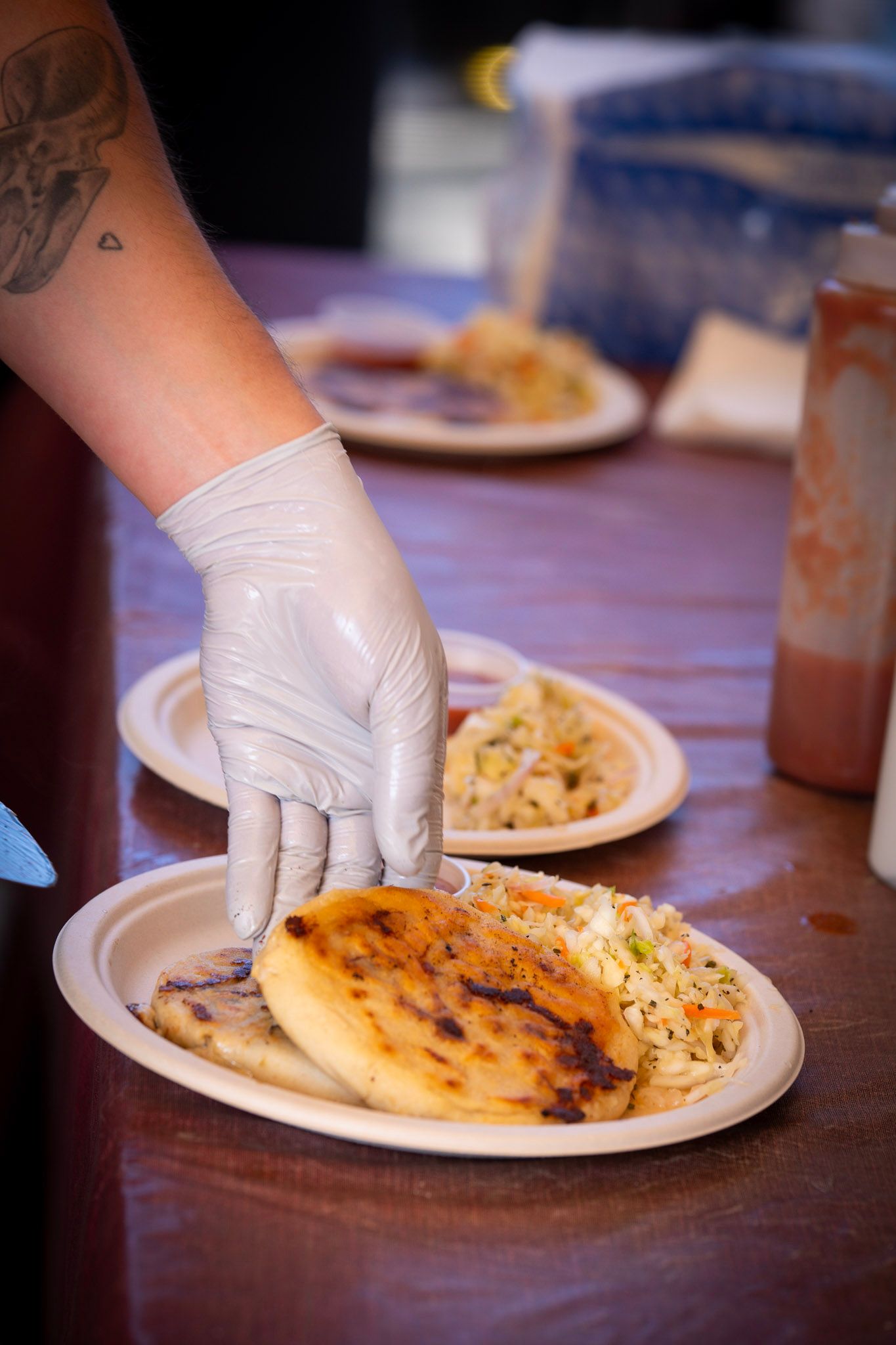 Person wearing a glove serving pupusas and slaw on a white plate at a food stand.
