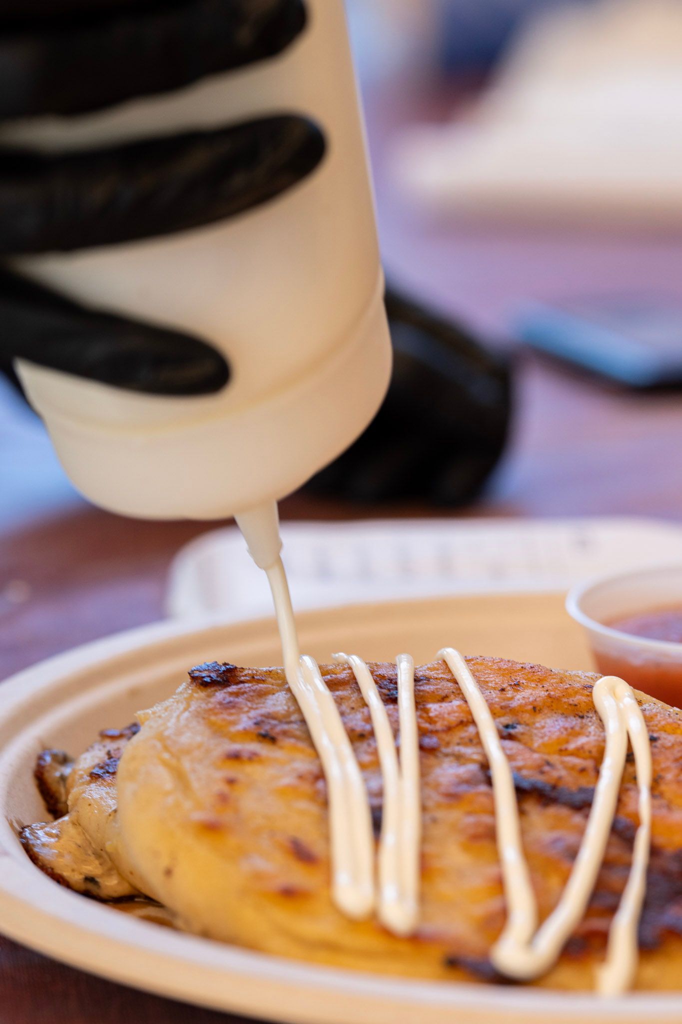 Hand in black glove squeezing white sauce onto grilled food on a plate, with a small sauce cup.