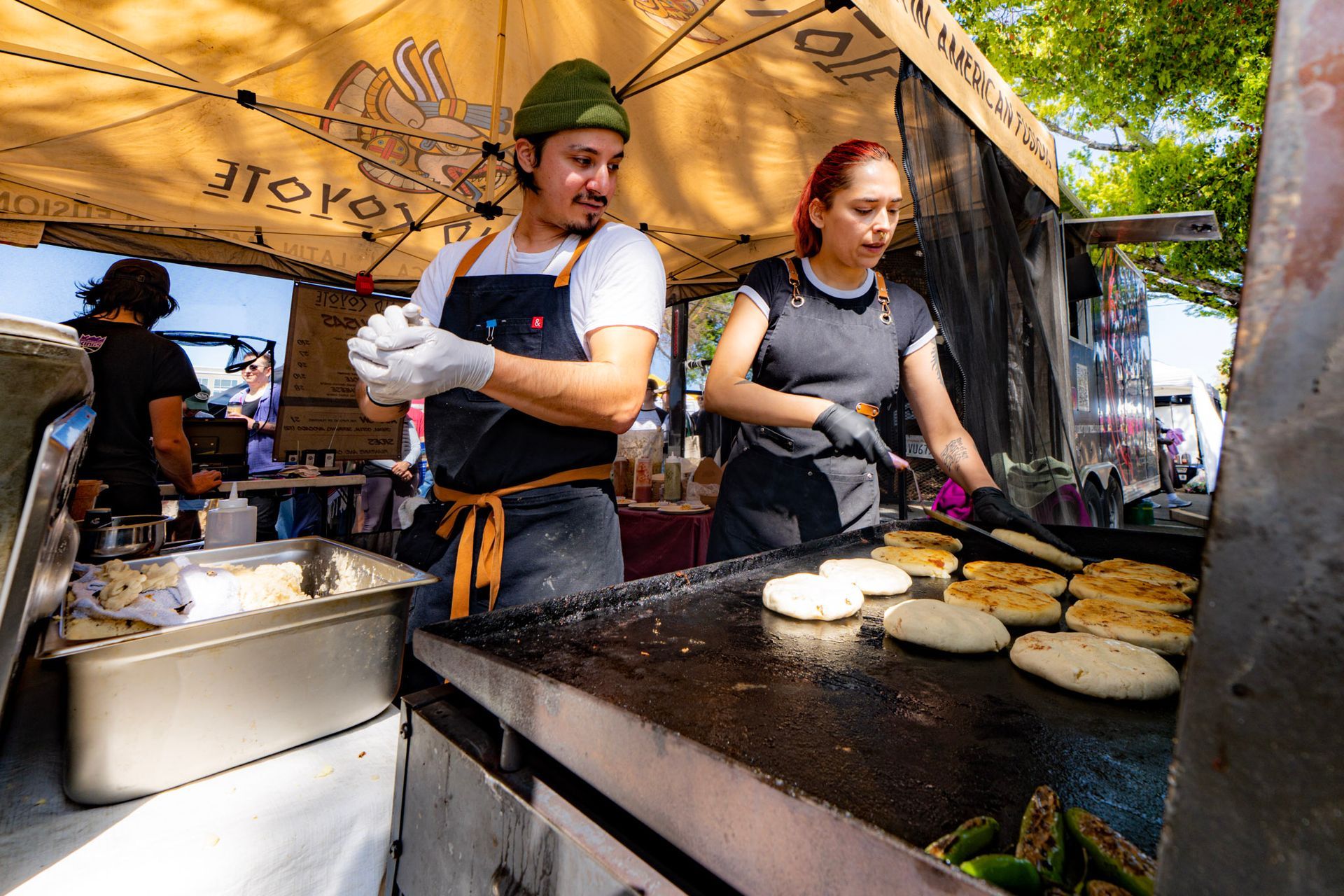 Two people cooking arepas at a food stall. Man in apron and hat prepping; woman flipping arepas. Sunny outdoor setting.