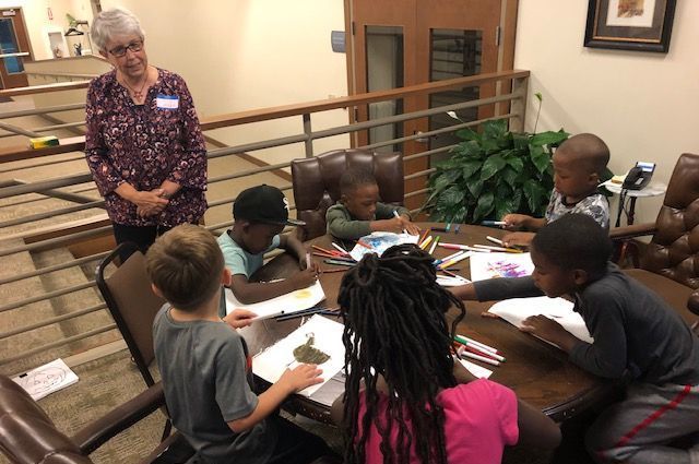 A group of children are sitting around a table with a woman standing behind them.
