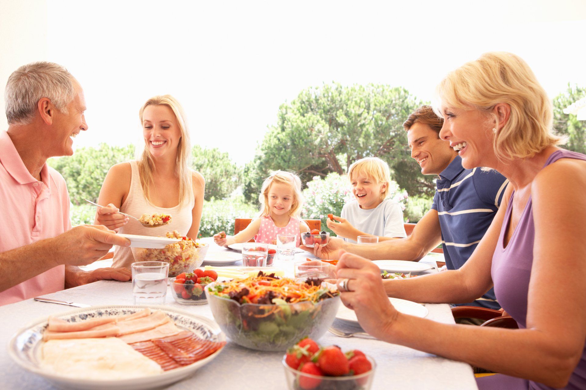 Famille déjeunant dehors, souriante. Salade, fruits et charcuterie sur la table.