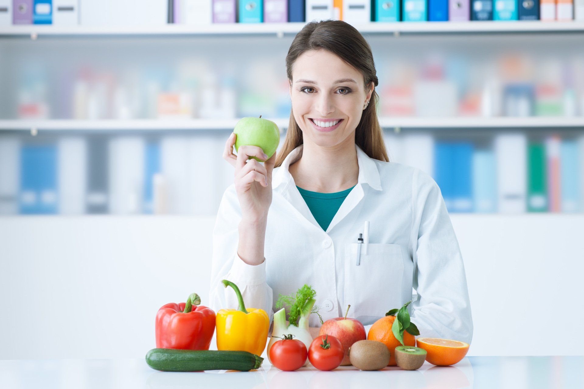 Femme en blouse de laboratoire souriante, tenant une pomme verte, entourée de fruits/légumes.