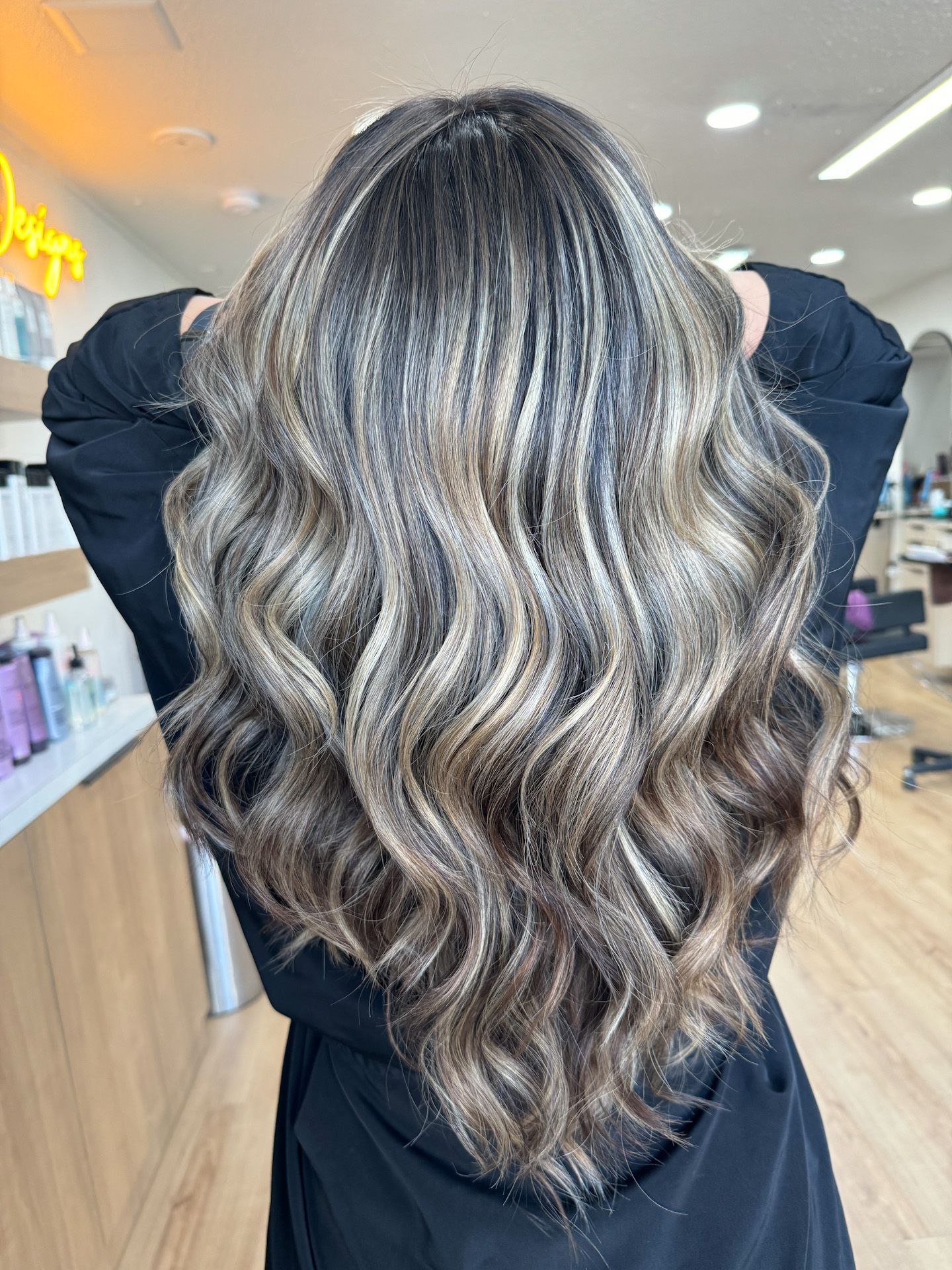 A woman with long wavy hair is standing in a salon with her hands behind her head.