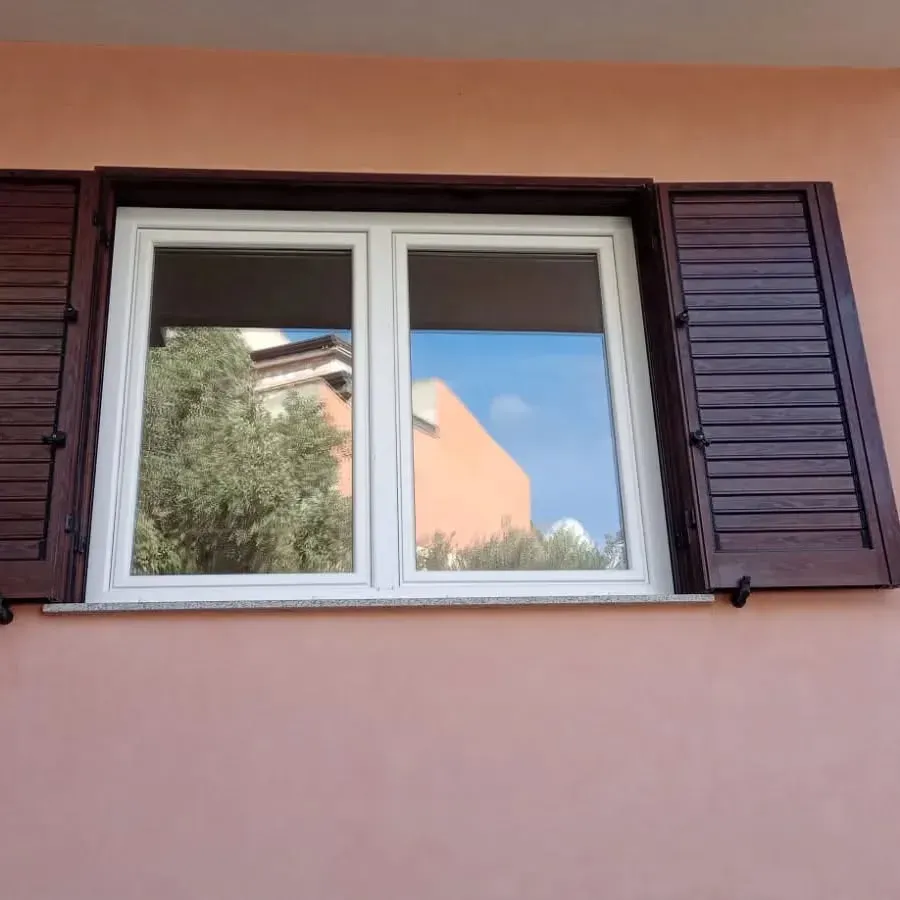 Finestra con cornice bianca, persiane marrone scuro, su cui si riflettono un albero e degli edifici contro una parete rosa.